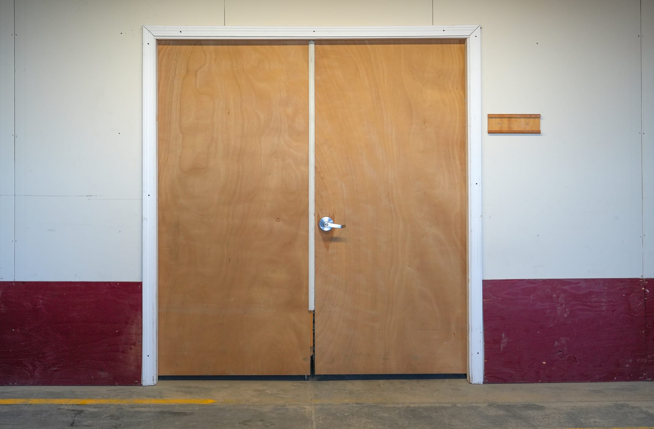 Double industrial wood doors with metal lever handle in a commercial warehouse setting, framed by white trim and surrounded by drywall and red lower wall paneling.