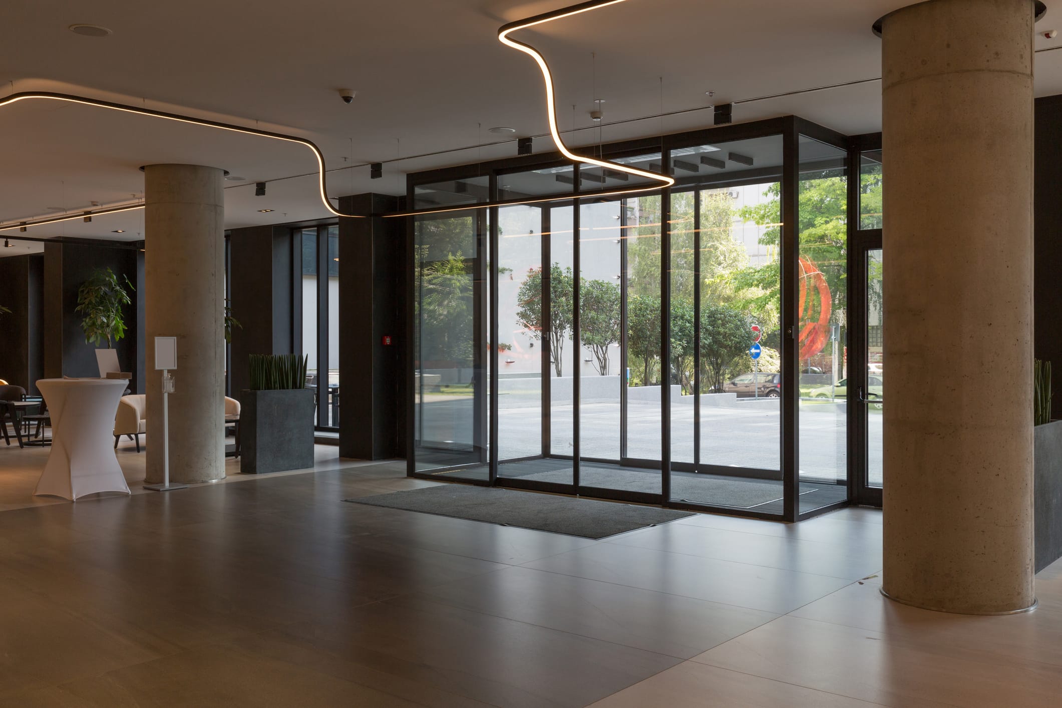 Modern commercial building entrance with large glass sliding doors and a clean, sleek metal frame, surrounded by reflective tile flooring and indoor greenery.