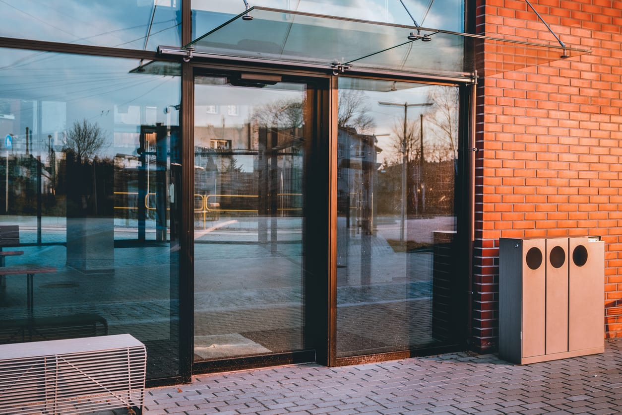 Modern black-framed glass storefront reflecting sunset light, adjacent to a red brick wall. The entrance features a glass canopy overhead and a paved walkway.