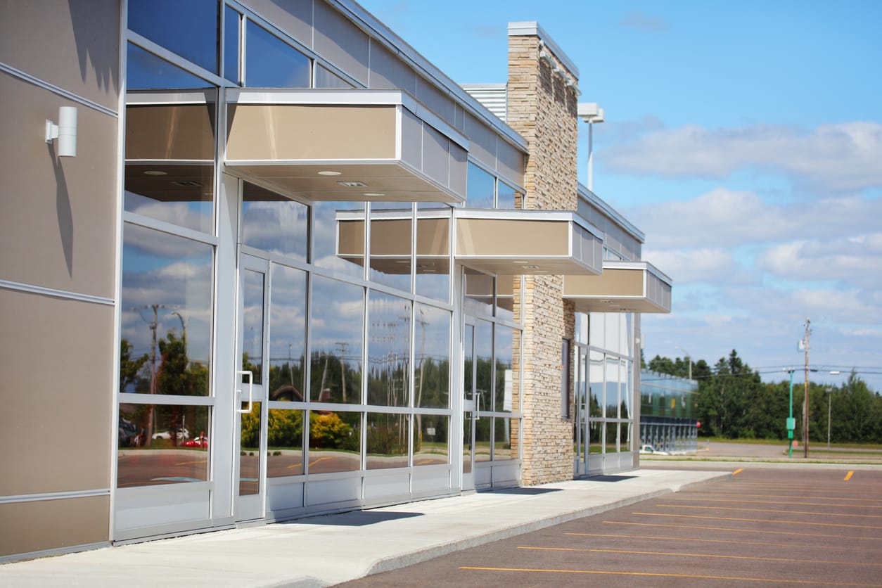 Modern retail strip mall featuring tan-colored awnings, large glass storefronts, and a stone accent wall. The sky is blue with scattered clouds and an empty parking lot is in the foreground.