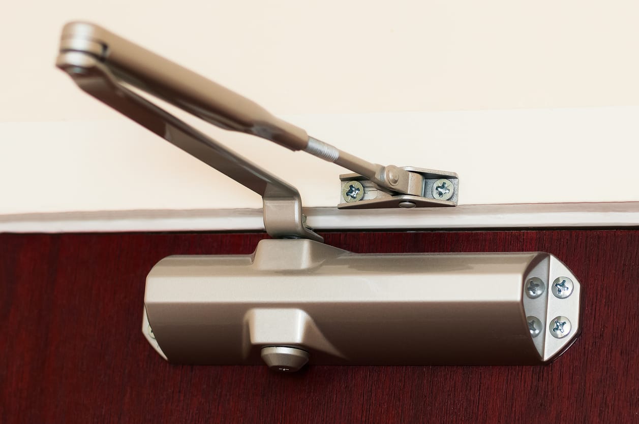 Close-up of a commercial door closer in a metallic finish, mounted on a wooden door to ensure smooth closing.