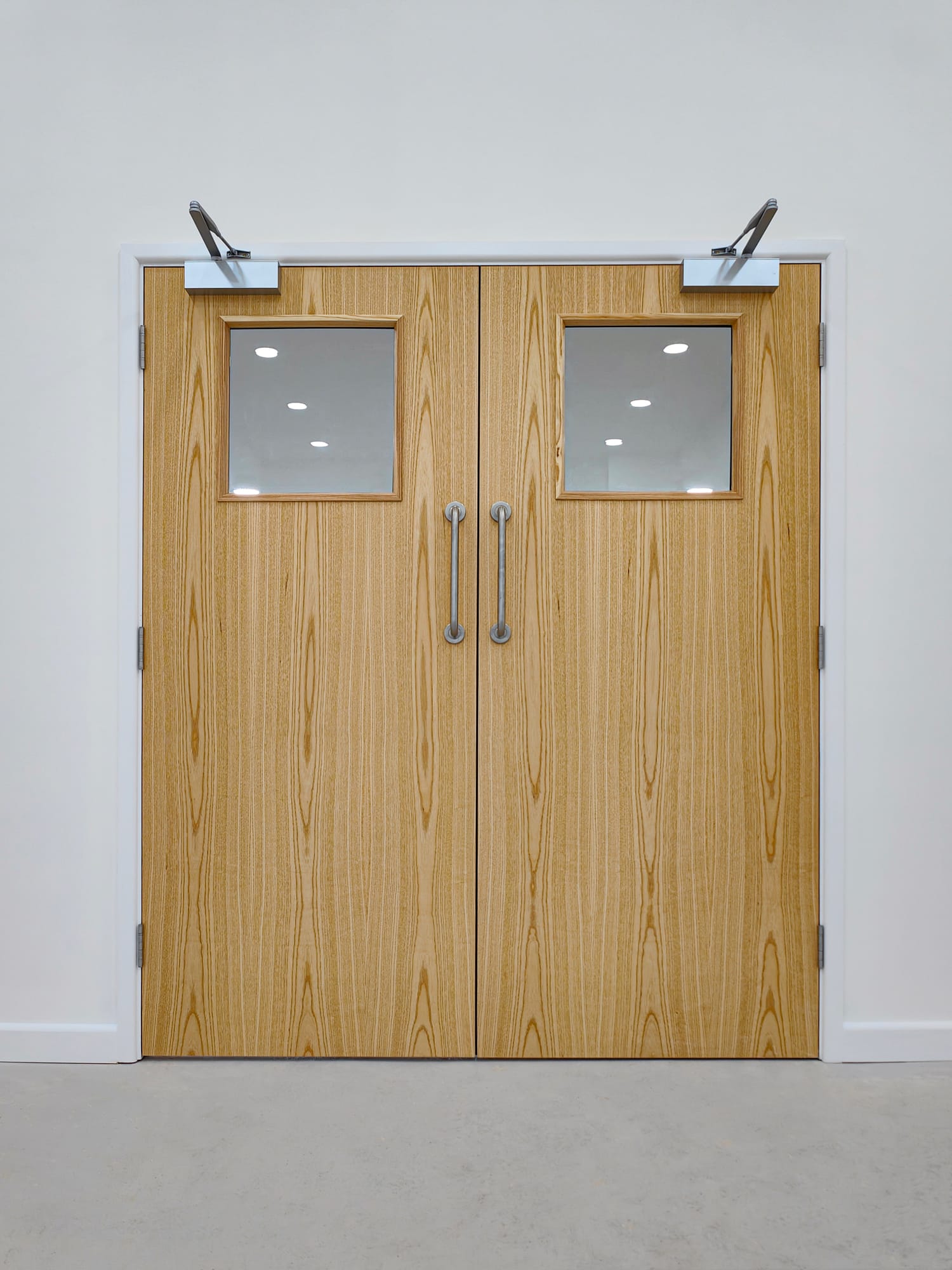 Light wood double commercial doors with square glass vision panels and metal push-pull hardware, set in a white wall with overhead door closers.