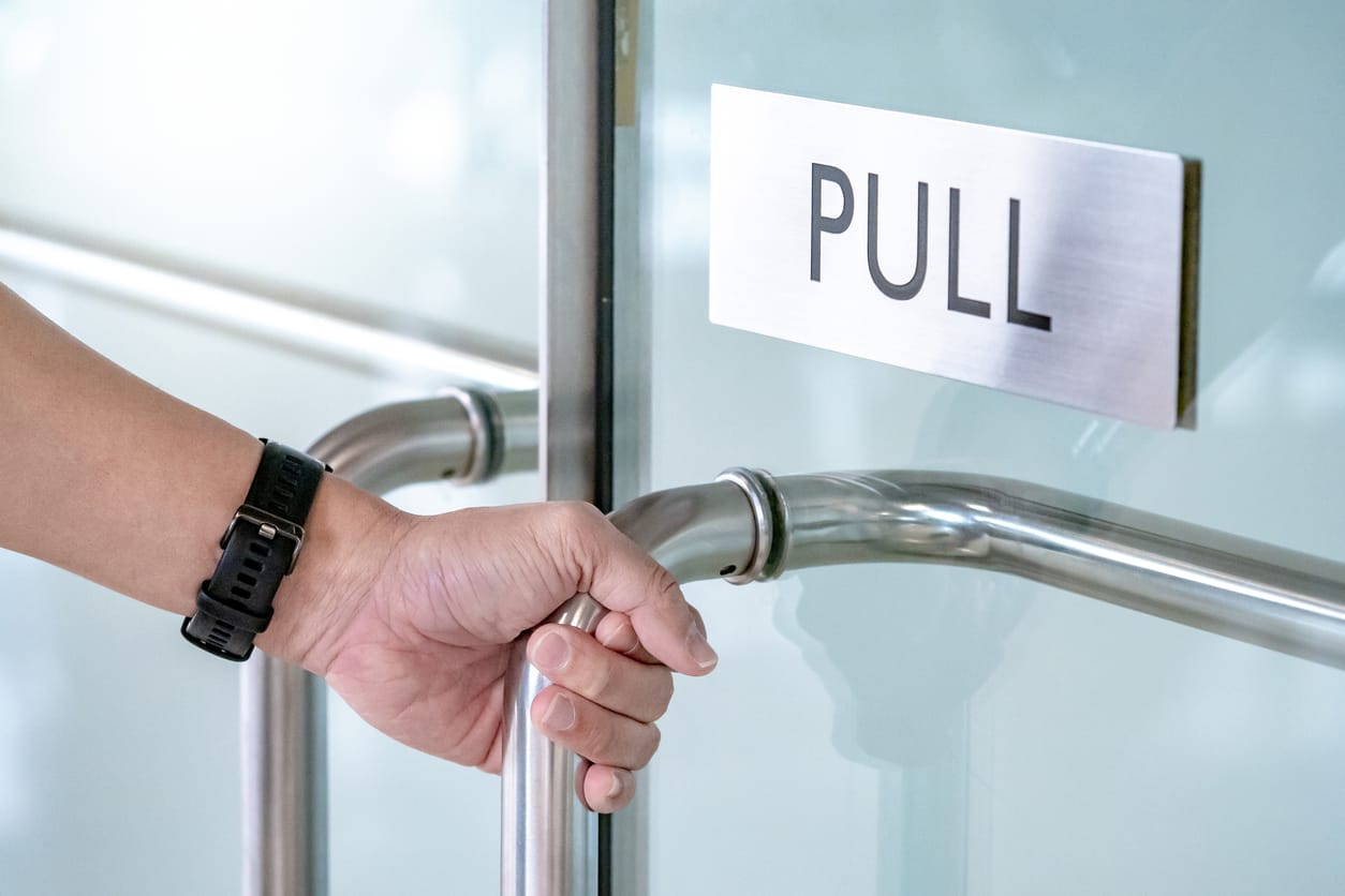 Close-up of a person’s hand gripping a stainless steel pull handle on a commercial door, with a brushed metal plate engraved with 'PULL' on a glass panel background.