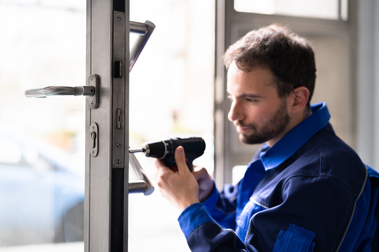 Close-up of a maintenance technician in blue overalls using a cordless drill to perform commercial door repair, focusing on the door handle and lock mechanism.