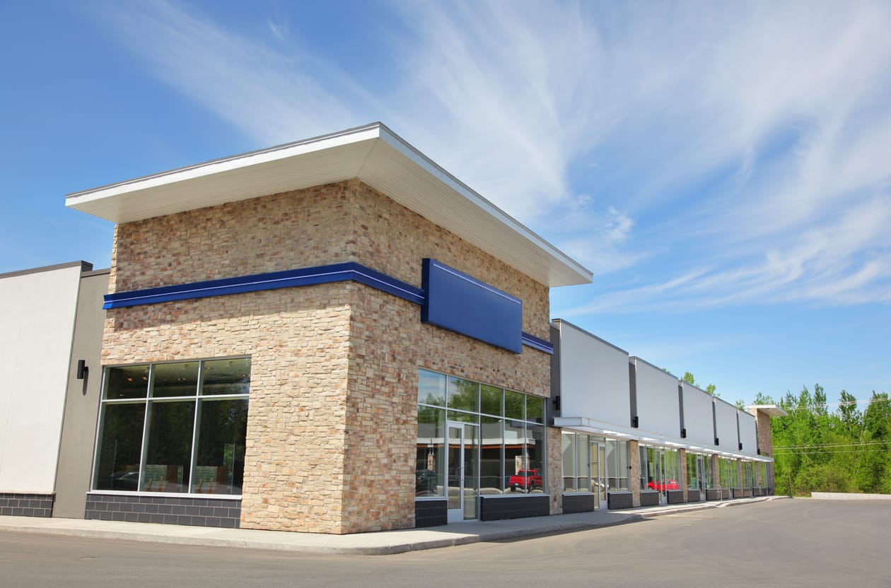 Modern commercial strip mall featuring a prominent corner unit with a tan brick facade and a blank blue sign. Large glass storefront windows. The surroundings include paved roads and clear skies.