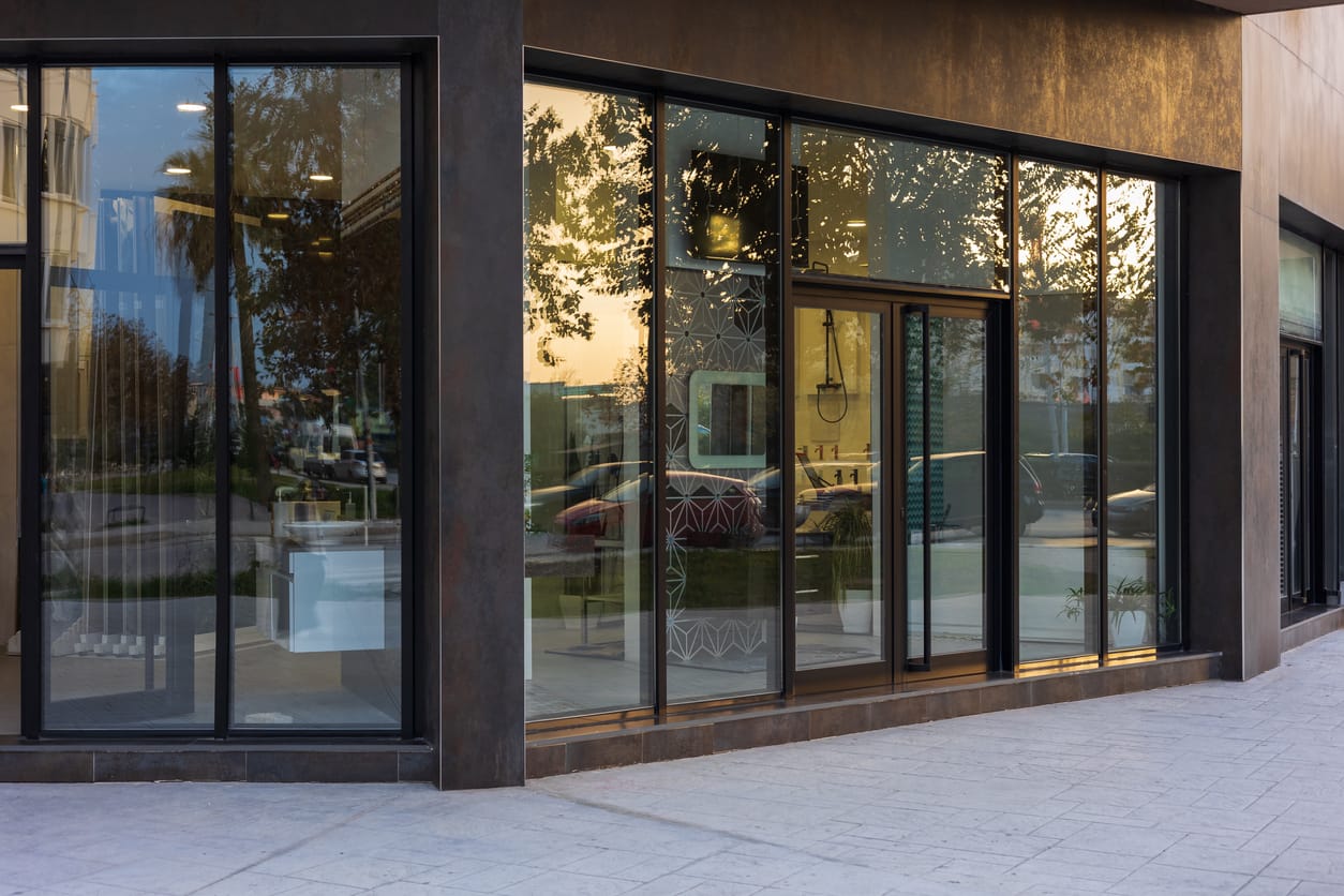 Modern furniture store with large glass windows reflecting sunset and trees. The dark metal frame of the storefront gives a contemporary look.