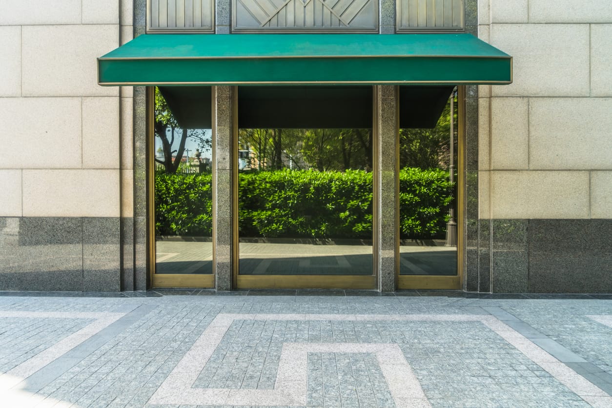 Modern glass storefront with reflective green glass panels, a large green awning, and a polished stone facade. The sidewalk features a decorative tile pattern.