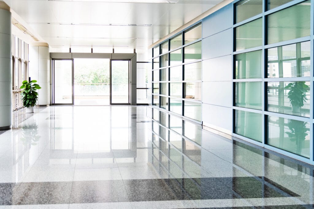 Modern office building lobby with large windows and commercial doors. The space is bright with reflective flooring and potted plants.