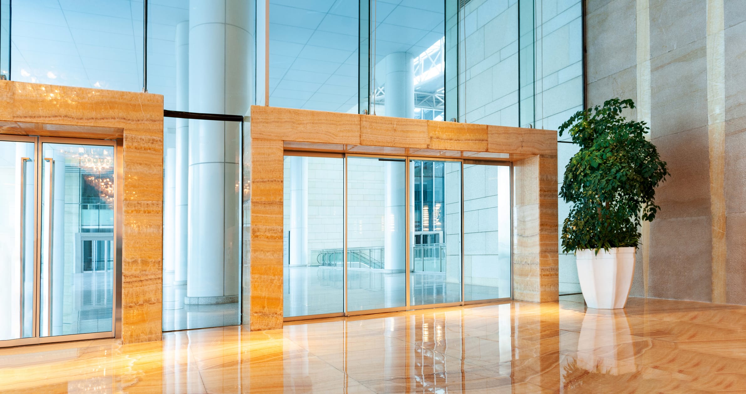 Spacious office lobby featuring wide automatic sliding glass doors framed in light stone, with a high ceiling and polished marble floors reflecting natural light.