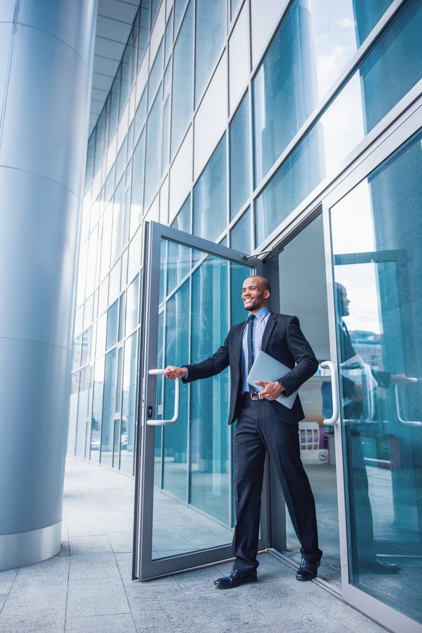 Confident businessman holding a laptop, stepping out of a modern commercial building through a glass commercial door with large windows and a sleek design.