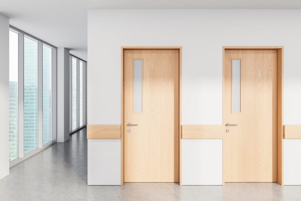 Two modern light wood office doors with narrow vertical glass windows, set in a white wall within a bright commercial hallway. Large floor-to-ceiling windows on the left reveal a cityscape, filling the space with natural light and highlighting the clean, professional office design.