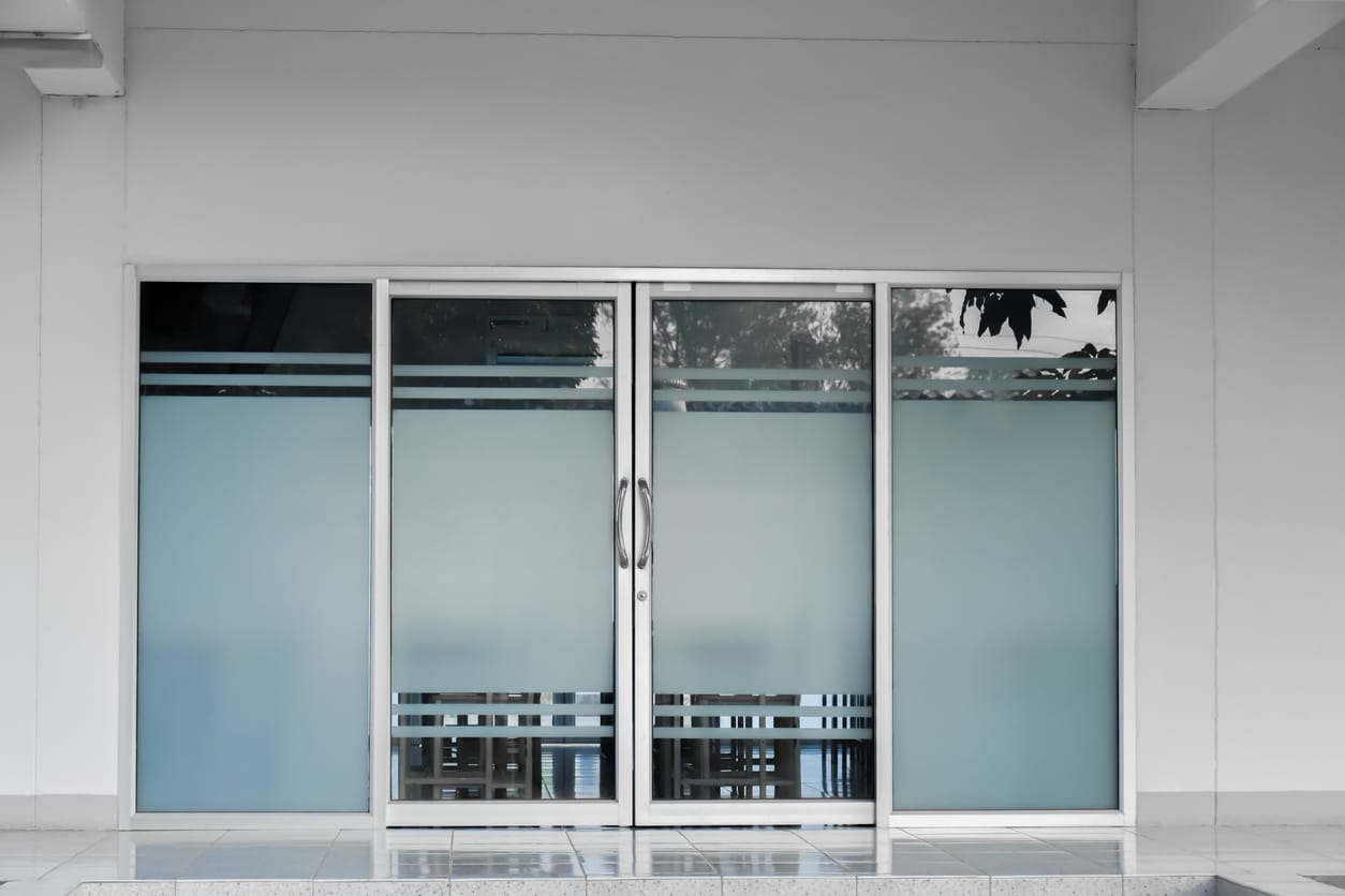 Modern glass doors with frosted panels and metal handles, installed in a white-walled commercial building. Reflections of trees and surroundings are visible in the upper panels.