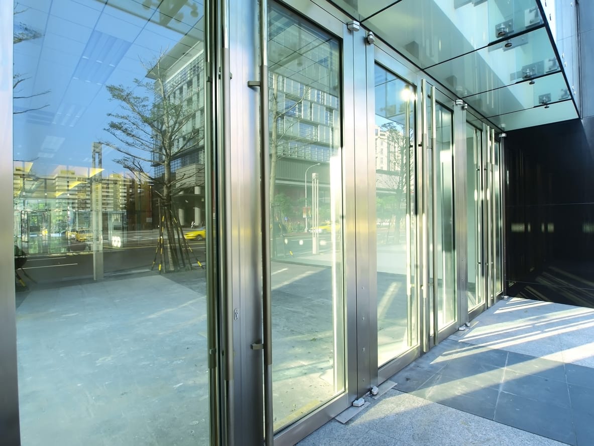 Modern commercial building featuring floor-to-ceiling glass storefront windows and steel frames. The entrance area reflects a vibrant cityscape with trees and urban buildings in the background.