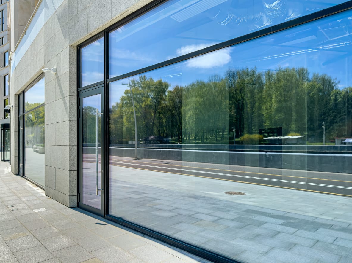 Modern commercial building exterior featuring large black framed glass windows and doors with a granite stone facade. Trees are reflected in the glass, and the street has a tiled sidewalk.