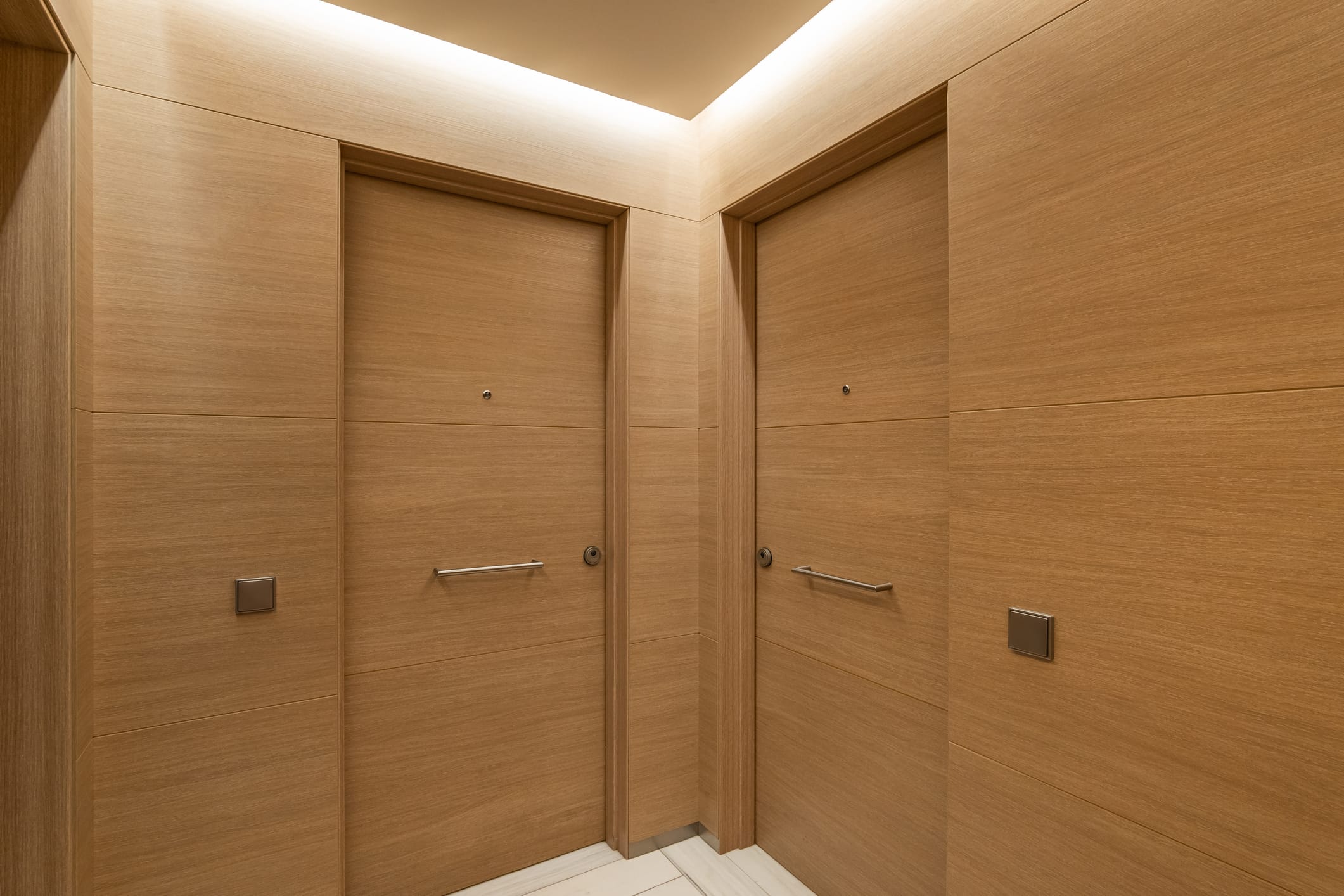 Two minimalist wooden apartment doors meet at a corner in a modern hallway. The space features uniform wood paneling, discreet door viewers, and sleek metal handles, enhanced by warm recessed lighting.
