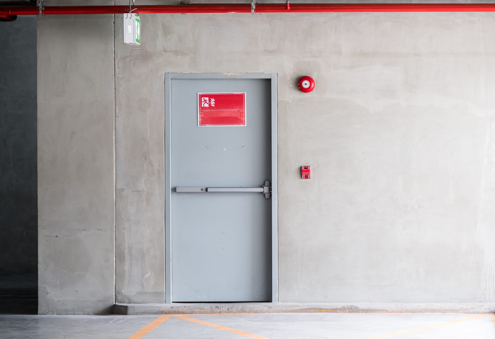 Thanks for sharing the image. It shows a metal fire exit door in what looks like a parking garage or industrial setting. The door features a panic bar for emergency egress, a red fire exit sign, and nearby fire alarm components (manual pull station and alarm bell). There's also an exit sign above the door and red fire sprinkler piping on the ceiling.