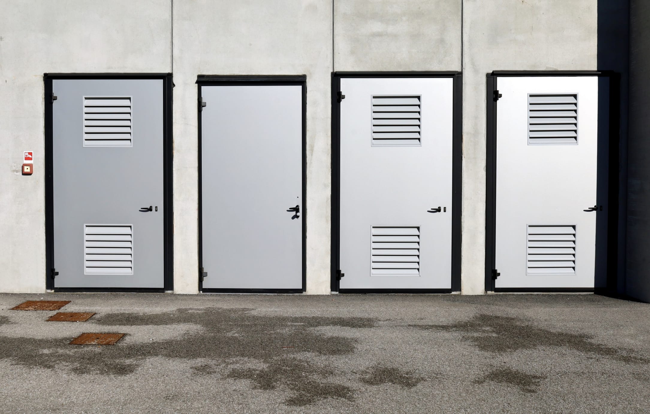 Four industrial metal doors on a concrete building exterior, three featuring built-in ventilation louvers, commonly used for utility or mechanical rooms.