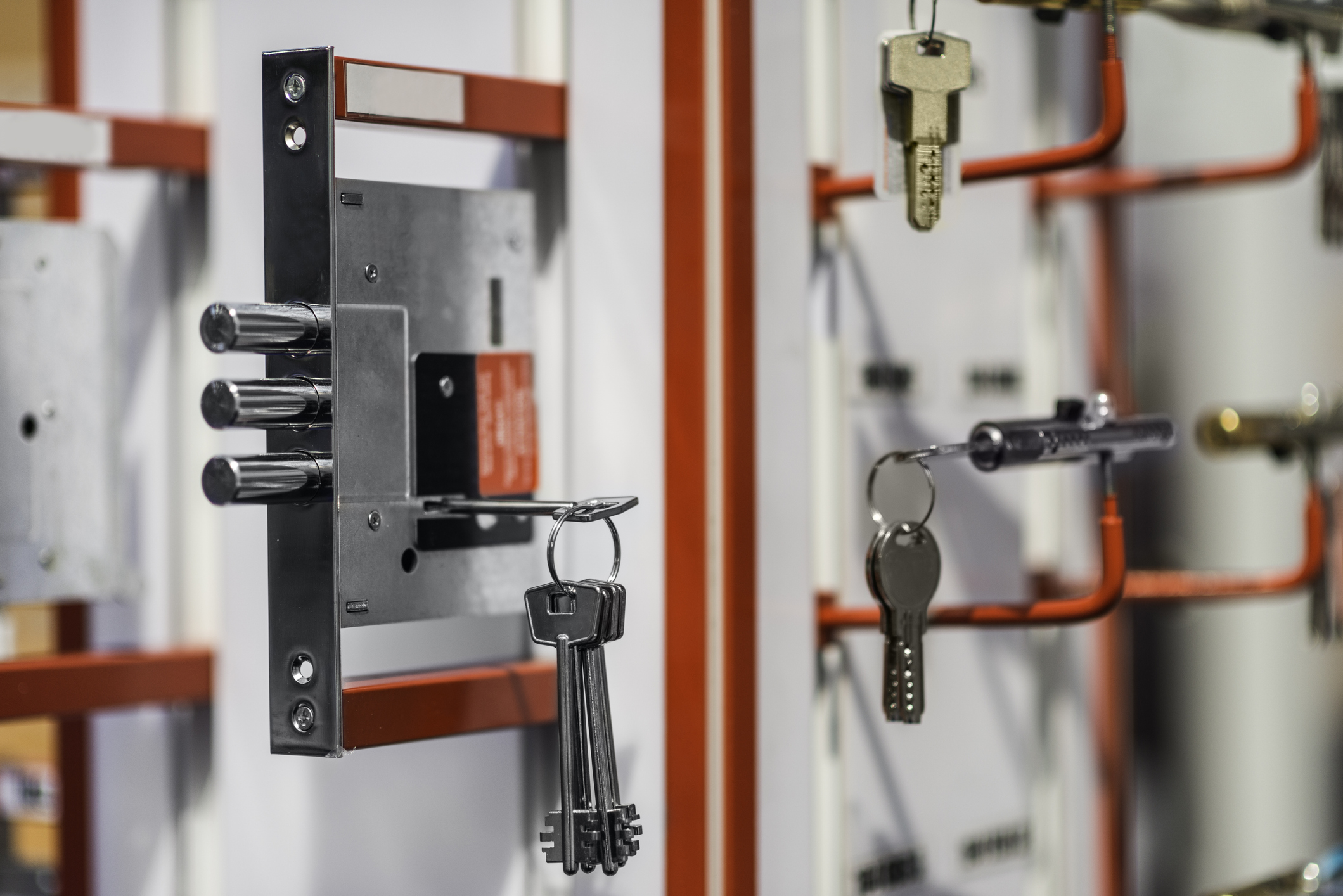 Close-up of a mortise cylinder lock with multiple keys inserted, displayed on a red and white wall rack with other lock mechanisms and key sets in the background.