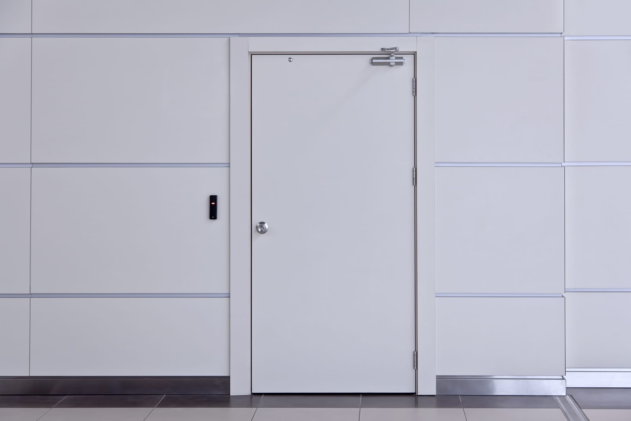 White commercial security door with metal frame, door closer, and access control reader mounted on the adjacent wall in a modern office building.